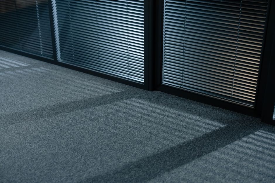 A long, narrow corridor in a commercial building with white walls and a suspended ceiling fitted with evenly spaced recessed lighting. On the left side, there are several closed black doorways, each w