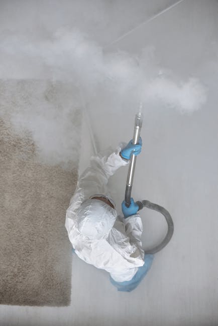 A professional cleaner, dressed in white protective coveralls and blue gloves, is performing steam or high-temperature sanitisation on a white tiled floor in a residential room, possibly a kitchen or bathroom. The cleaner is using a handheld steam cleaner with a long metal wand and a hose, emitting visible steam onto the surface. The room is well-lit with natural or bright artificial lighting, highlighting the clean, smooth tiles and the adjacent plain white wall. A beige textured mat partially covers the floor near the cleaned area. The scene depicts precise surface cleaning and hygiene maintenance, emphasizing deep cleaning processes as offered by Carpet Cleaning Pimlico for apartments such as Eccleston Square, ensuring hygienic and spotless living spaces.