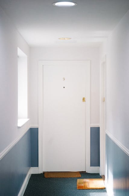 A bright, narrow hallway with white walls and a blue wainscot paneling running along the lower half, illuminated by natural light from a window on the left side. The flooring is covered with a dark carpet, and a small brown doormat is placed in front of a closed white door labeled with the number 9, featuring a brass handle and peephole. The ceiling is clean and smooth, with a recessed light fixture. The space appears well-maintained and free of dust or clutter, exemplifying a neat environment suitable for surface cleaning and domestic hygiene, as provided by Carpet Cleaning Pimlico.