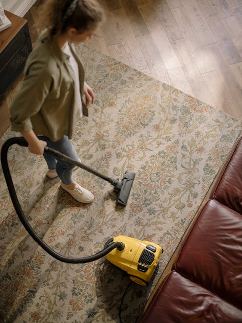 A woman in casual clothing is performing surface cleaning on a large, patterned area rug with a vacuum cleaner in a residential living room. The rug features intricate floral designs in muted colors, and is placed on a wooden floor. The living room has natural lighting, with part of a red leather sofa visible nearby. The scene depicts deep cleaning after flooding, with attention to hygiene and surface upkeep, which is typical of services provided by Carpet Cleaning Pimlico. The vacuum cleaner is yellow with a black hose, and the woman is focused on ensuring thorough sanitation of the rug, highlighting professional domestic cleaning procedures.
