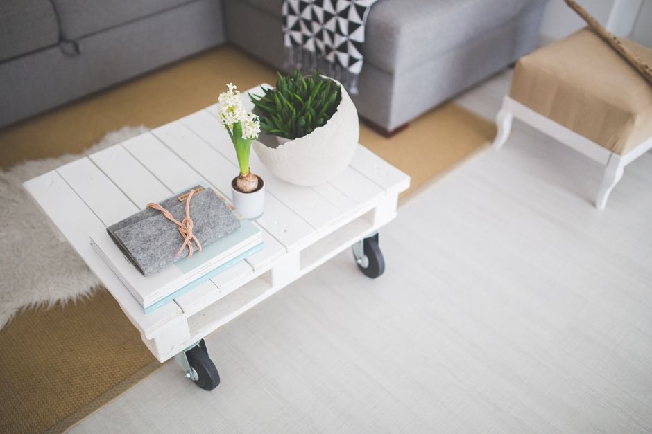 A white wooden coffee table with black caster wheels situated in a living room, featuring a small potted plant with green leaves and white flowers, a grey felt blanket tied with a light pink ribbon, a