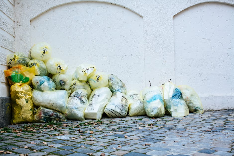 A row of large yellow and white refuse bags filled with bulky waste are piled along a cobblestone street, adjacent to a white brick and concrete wall. The bags contain discarded household items and are tied with black strings. The ground shows scattered leaves and debris, and the lighting suggests an outdoor setting during daylight. This image illustrates waste collection for bulky waste disposal, relevant to cleaning and maintenance services provided by Carpet Cleaning Pimlico in Pimlico, highlighting the importance of proper waste management and surface cleaning.