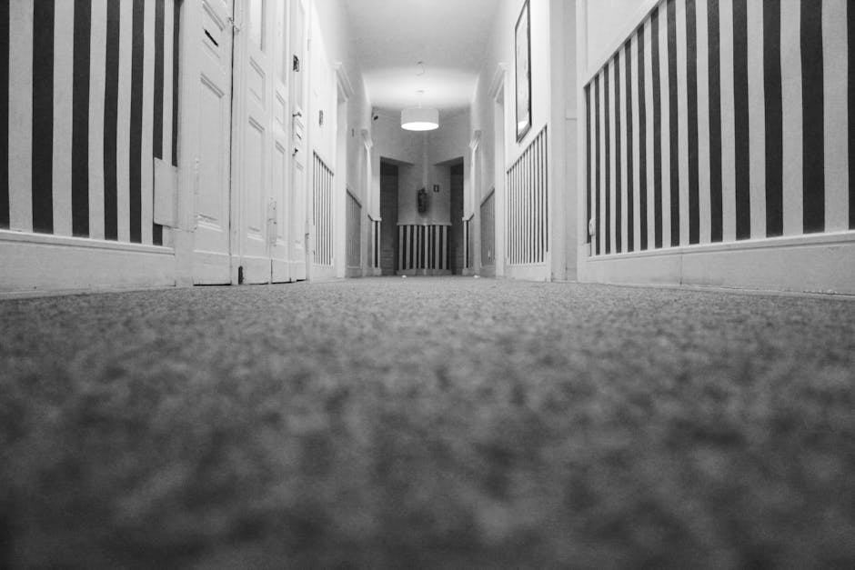 A black and white photograph of a hallway with a low-angle view, focusing on the carpeted floor which appears clean and well-maintained. The hallway has white wooden wainscoting on the walls, with vertical striped wallpaper above. Several closed white doors line each side of the corridor, leading to rooms, and a round ceiling light fixture illuminates the space. The layout is symmetrical, with a glimpse of an open doorway at the far end. Carpet Cleaning Pimlico has expertise in surface cleaning and domestic cleaning, ensuring hygiene and maintenance for residential spaces like Pimlico basement flats, as highlighted in their webpage about mould and damp carpet fixes.