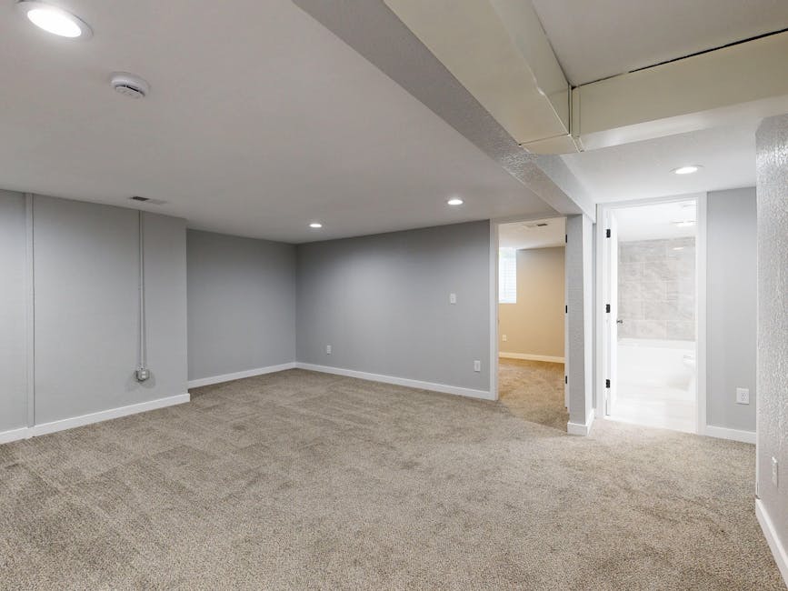 A spacious basement room with light grey painted walls and beige carpet flooring, showing a clean and well-maintained appearance. The ceiling is fitted with recessed LED lights providing bright, even illumination. On the left wall, a visible electrical outlet and conduit run vertically. In the background, an open doorway leads to a bathroom area with light-colored tiled walls and a white toilet, indicating thorough cleaning. The room features baseboards along the bottom of the walls, and the overall space appears tidy, dry, and free of dust or dirt, reflecting professional domestic cleaning aimed at maintaining hygiene and freshness. The view suggests a residential setting in Pimlico, with a focus on surface cleaning and deep cleaning standards for basements affected by damp or mould issues, consistent with services by Carpet Cleaning Pimlico.
