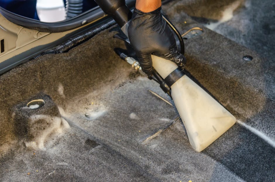 Close-up of a person's hand wearing a black glove, operating a cleaning device with a white plastic spray bottle and a hose attachment, used for deep cleaning and surface sanitisation of a dark grey carpeted floor in a commercial or domestic setting. The scene includes visible foam and liquid residue on the carpet, indicating active cleaning or rinsing. The background shows part of a metallic and plastic component, possibly from cleaning equipment or furniture. The environment appears well-lit, highlighting the cleanliness process overseen by Carpet Cleaning Pimlico, as part of their domestic cleaning services focused on surface and deep cleaning.
