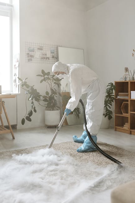 Close-up of a person's hand wearing a black glove, operating a cleaning device with a white plastic spray bottle and a hose attachment, used for deep cleaning and surface sanitisation of a dark grey carpeted floor in a commercial or domestic setting. The scene includes visible foam and liquid residue on the carpet, indicating active cleaning or rinsing. The background shows part of a metallic and plastic component, possibly from cleaning equipment or furniture. The environment appears well-lit, highlighting the cleanliness process overseen by Carpet Cleaning Pimlico, as part of their domestic cleaning services focused on surface and deep cleaning.