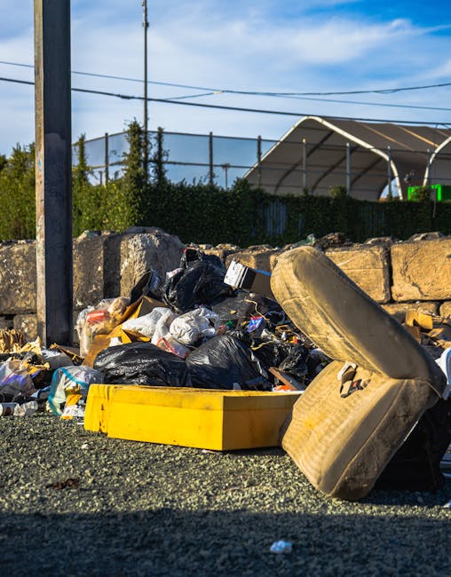 A street scene showing a pile of mixed waste including black plastic rubbish bags, a yellow plastic container, a large worn-out beige upholstered chair with visible dirt and stains, and some discarded